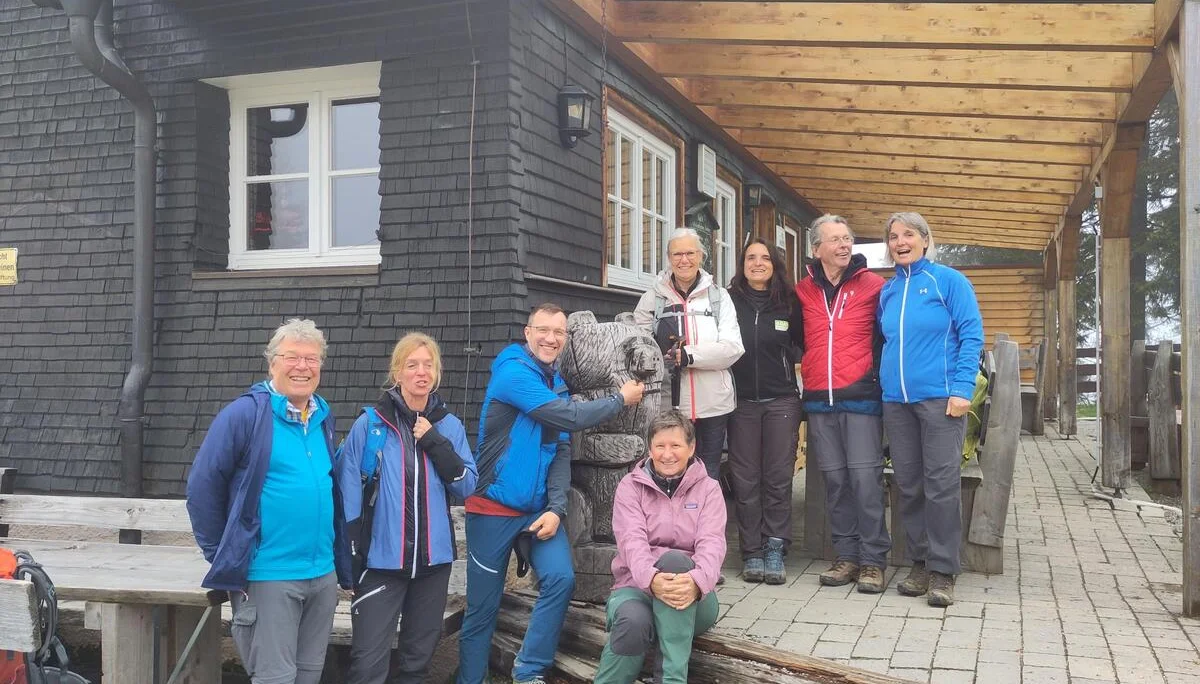 Acht Wanderer in Outdoor-Bekleidung vor einem Holzhütte stehen auf einer Veranda, einer umarmt eine Bärenfigur aus Holz | © DAV Sektion Geltendorf