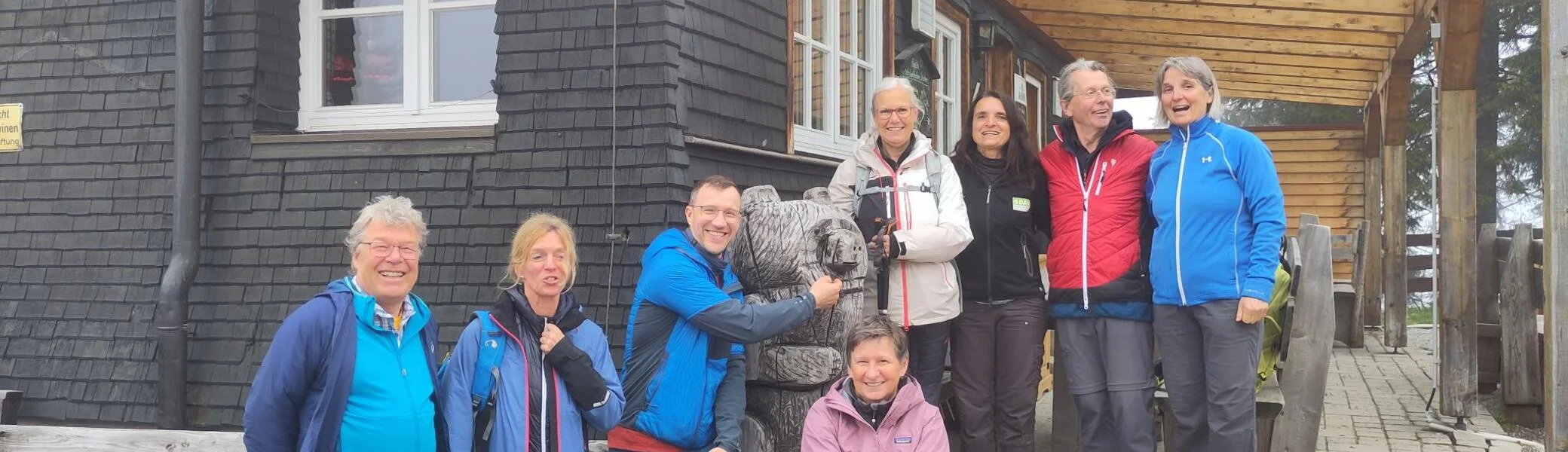 Acht Wanderer in Outdoor-Bekleidung vor einem Holzhütte stehen auf einer Veranda, einer umarmt eine Bärenfigur aus Holz | © DAV Sektion Geltendorf