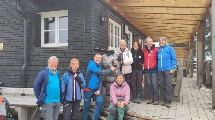 Acht Wanderer in Outdoor-Bekleidung vor einem Holzhütte stehen auf einer Veranda, einer umarmt eine Bärenfigur aus Holz | © DAV Sektion Geltendorf