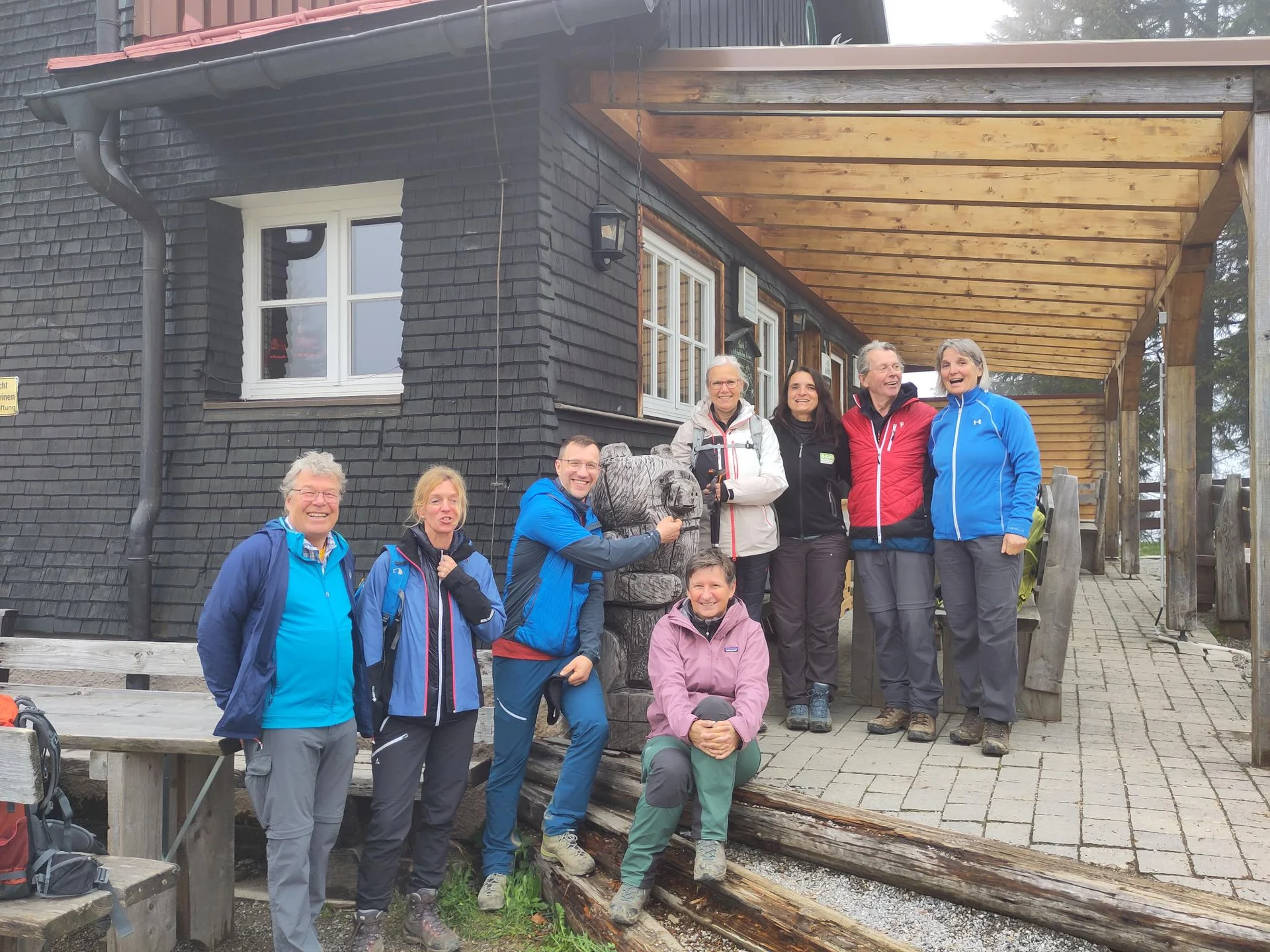 Acht Wanderer in Outdoor-Bekleidung vor einem Holzhütte stehen auf einer Veranda, einer umarmt eine Bärenfigur aus Holz | © DAV Sektion Geltendorf
