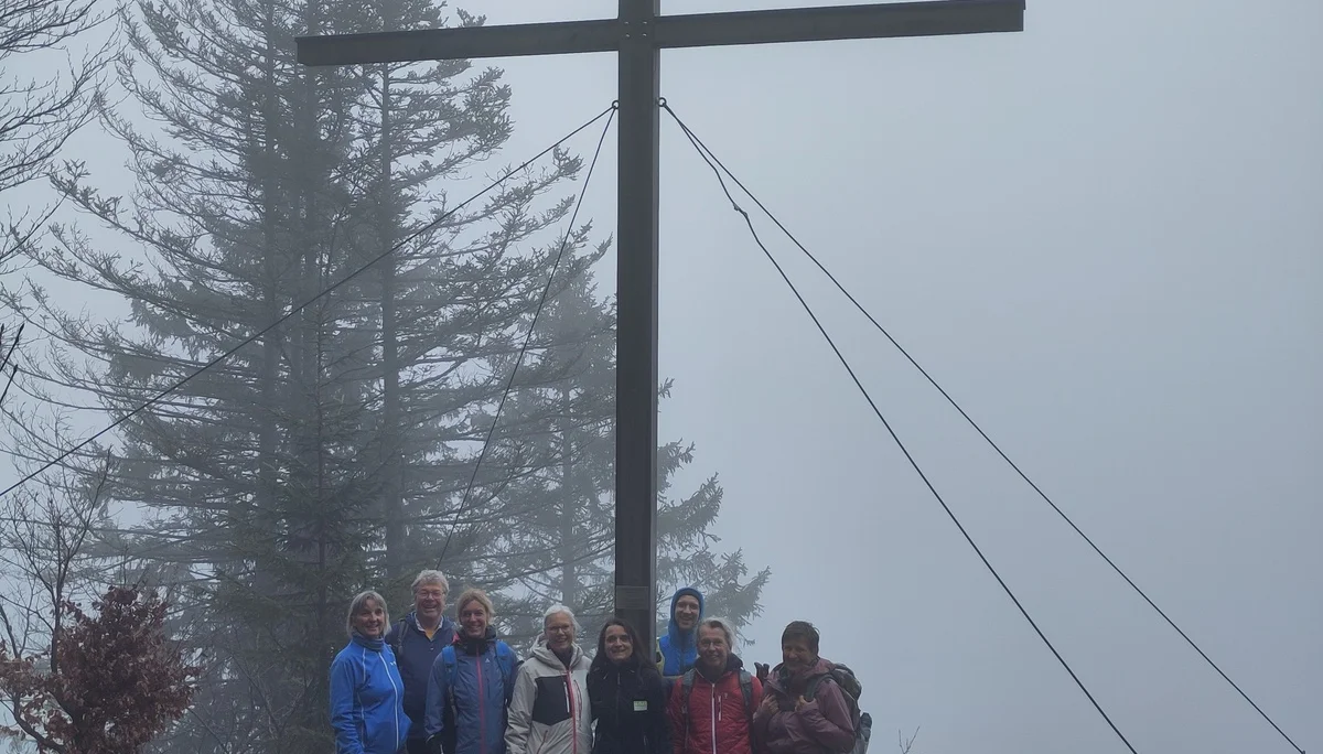 Gruppe von acht Personen steht neben einem Gipfelkreuz auf dem Gschwender Horn mit Bänken und Bäumen im Hintergrund | © DAV Sektion Geltendorf