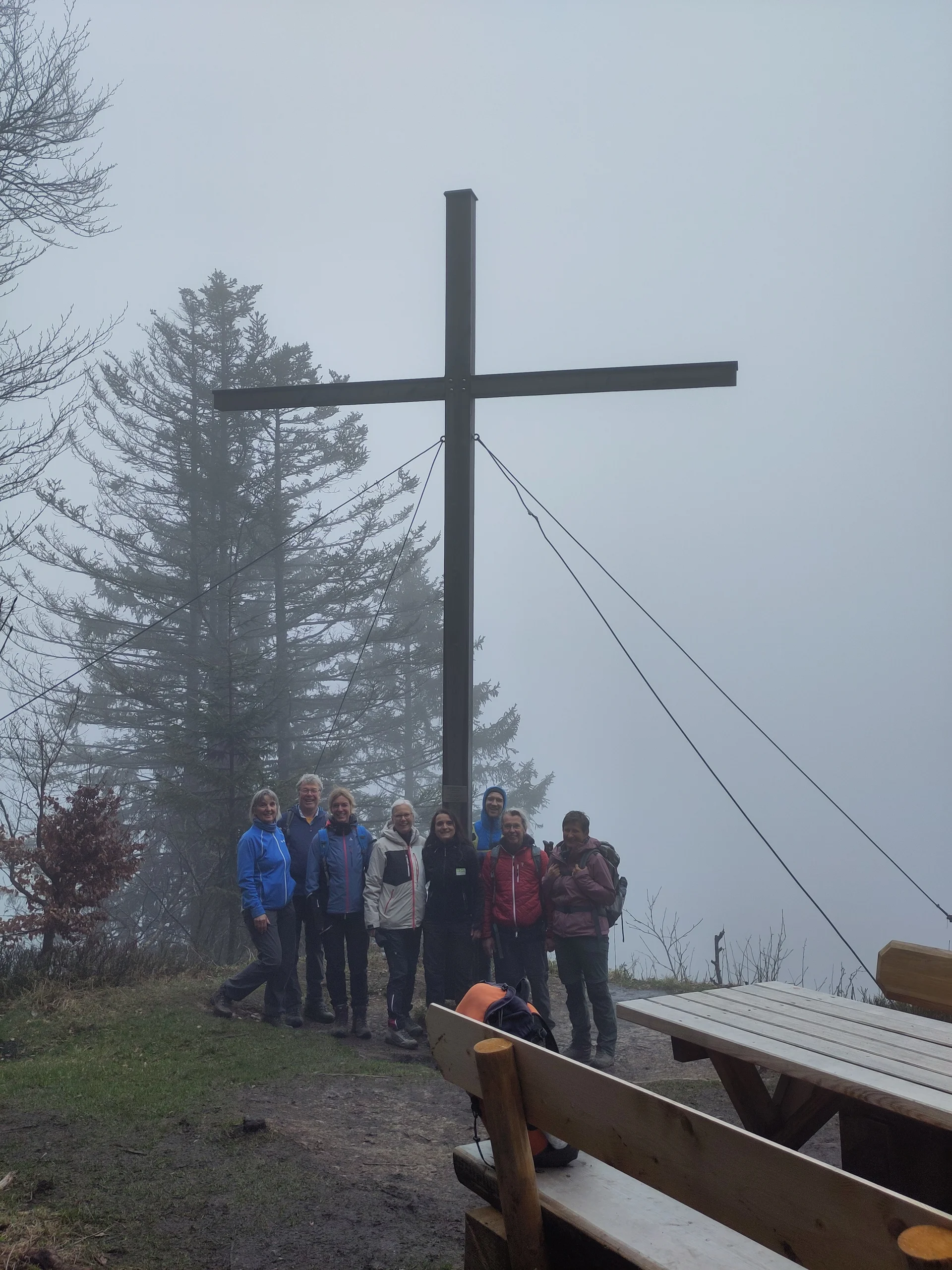 Gruppe von acht Personen steht neben einem Gipfelkreuz auf dem Gschwender Horn mit Bänken und Bäumen im Hintergrund | © DAV Sektion Geltendorf