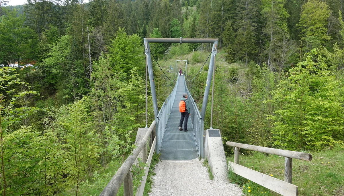 Person mit orangefarbenem Rucksack auf einer schmalen Hängebrücke über einem bewaldeten Tal | © Christoph Maier