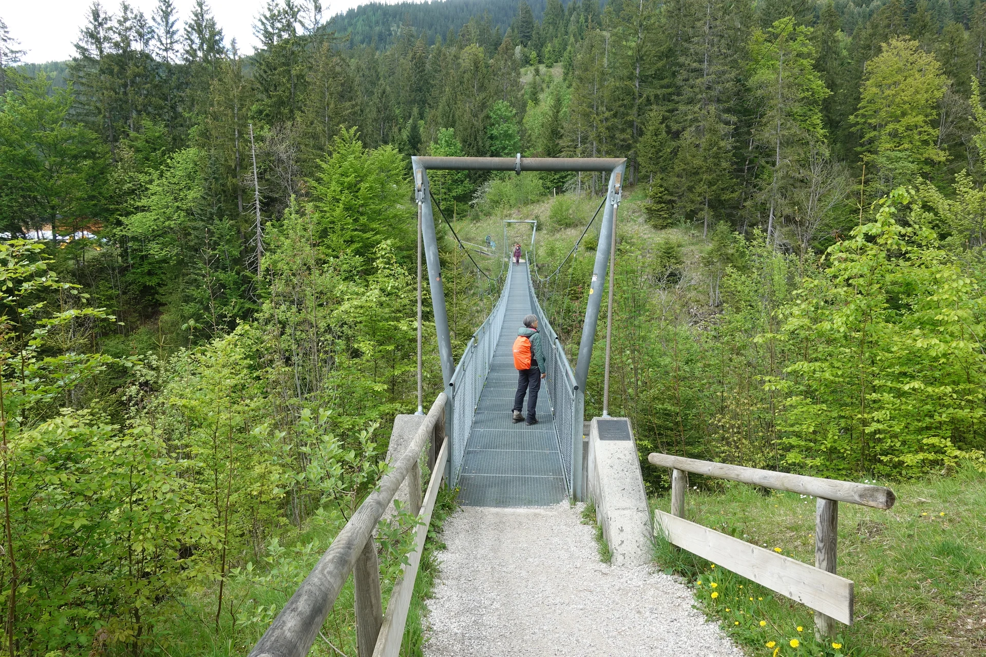 Person mit orangefarbenem Rucksack auf einer schmalen Hängebrücke über einem bewaldeten Tal | © Christoph Maier