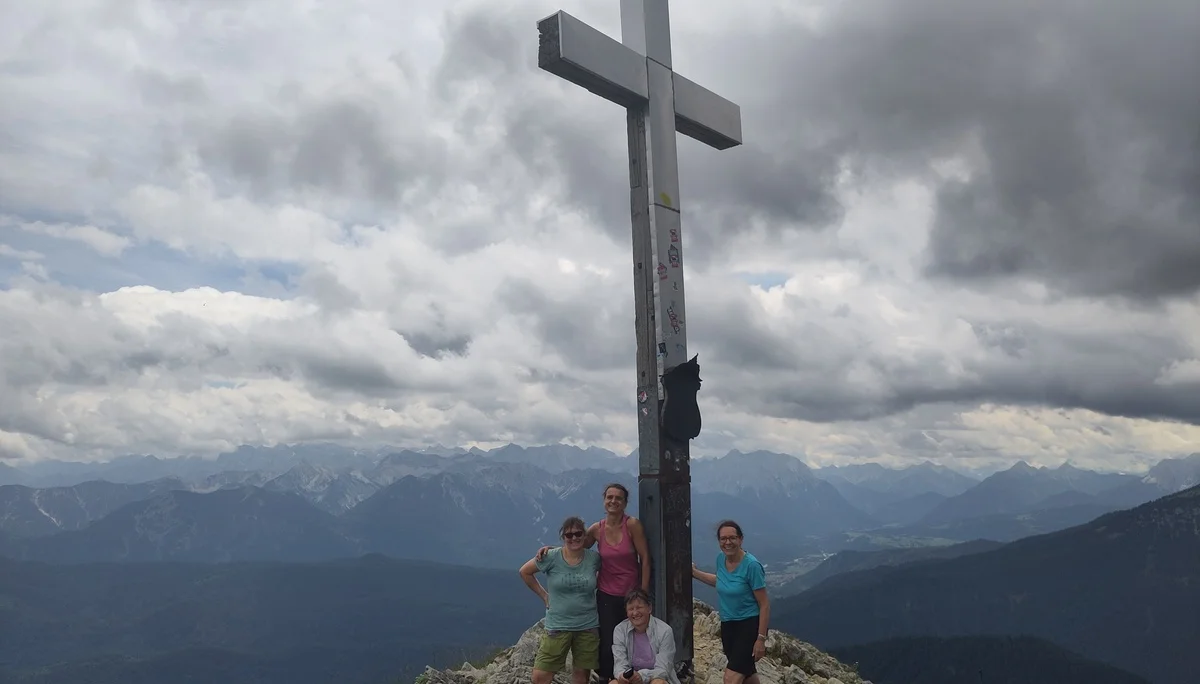 Vier Personen stehen und sitzen an einem Gipfelkreuz auf einem Berg mit bewölktem Himmel und Bergpanorama | © DAV Sektion Geltendorf
