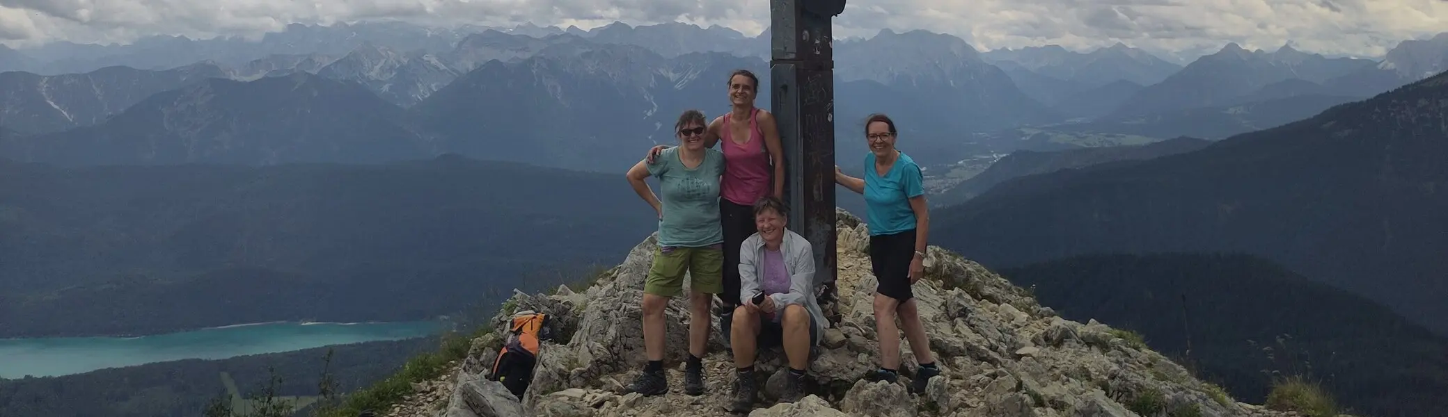 Vier Personen stehen und sitzen an einem Gipfelkreuz auf einem Berg mit bewölktem Himmel und Bergpanorama | © DAV Sektion Geltendorf