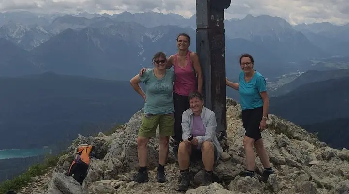 Vier Personen stehen und sitzen an einem Gipfelkreuz auf einem Berg mit bewölktem Himmel und Bergpanorama | © DAV Sektion Geltendorf