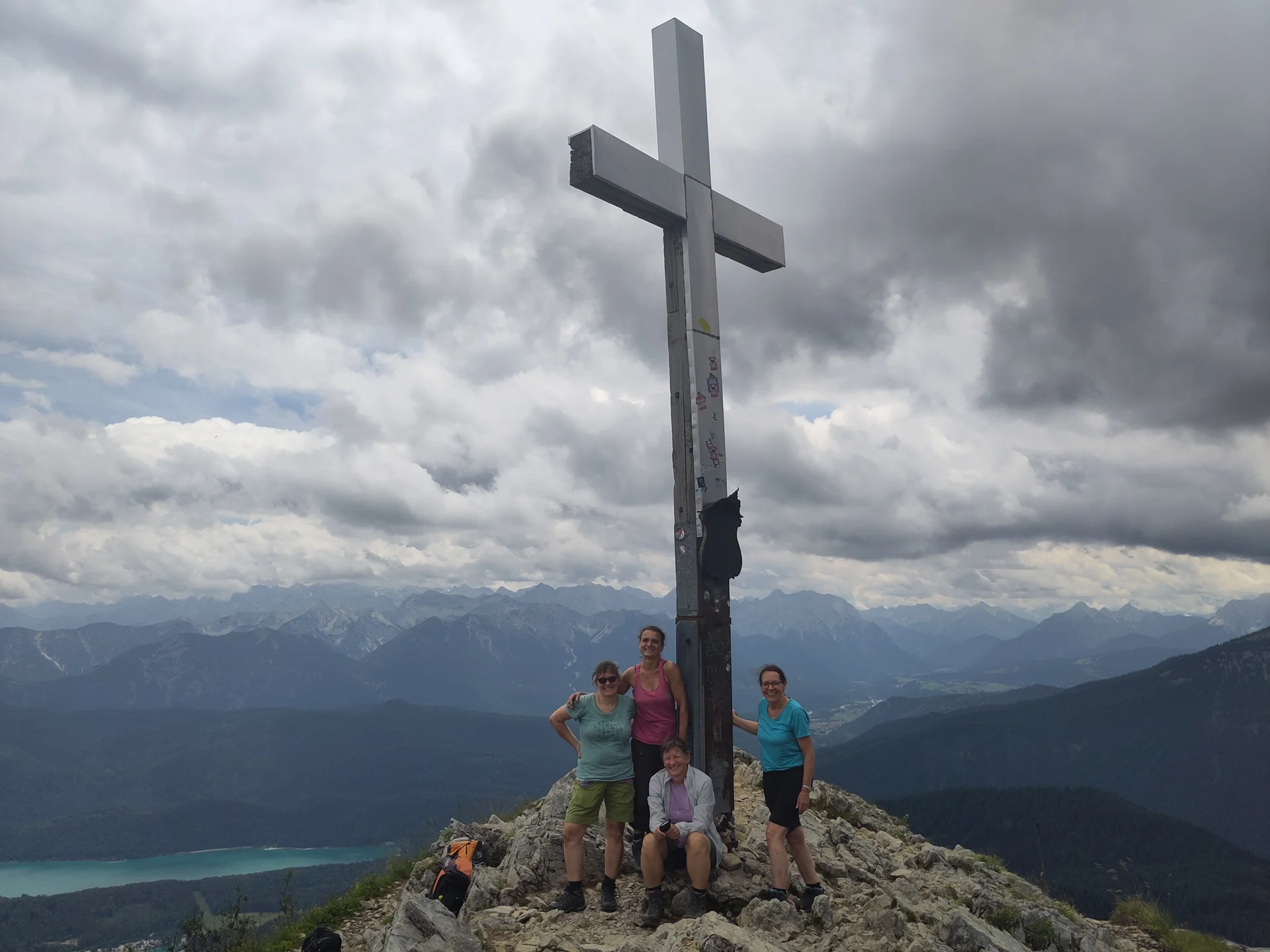 Vier Personen stehen und sitzen an einem Gipfelkreuz auf einem Berg mit bewölktem Himmel und Bergpanorama | © Sylvia Summerer