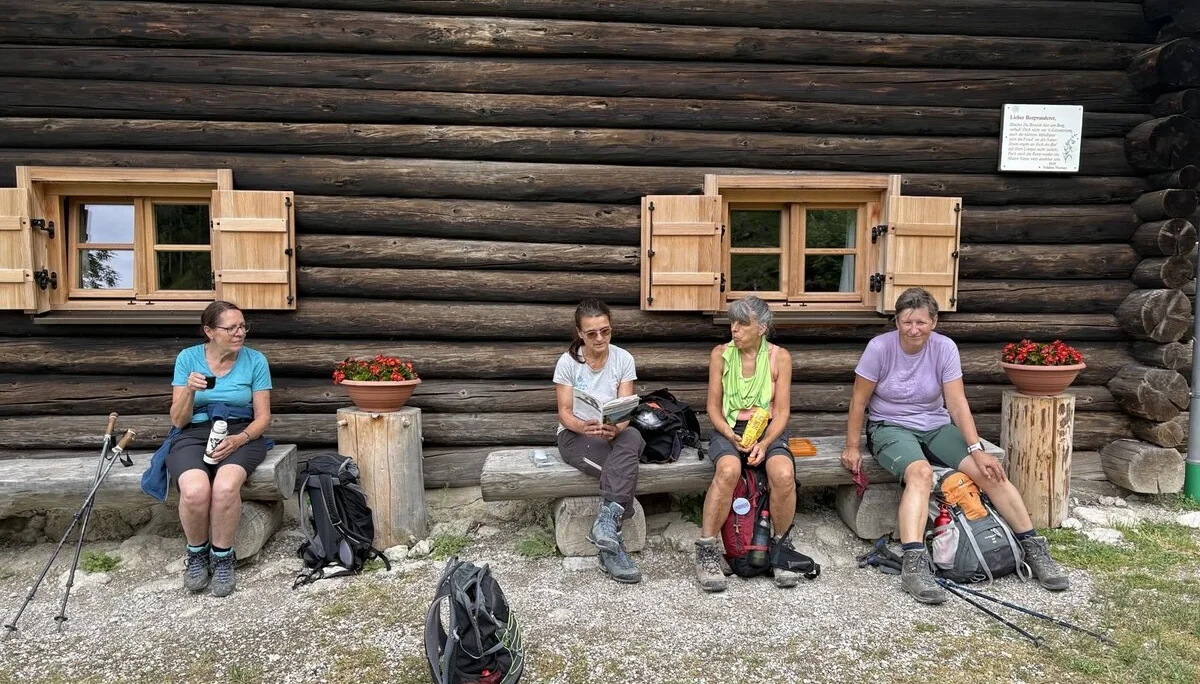Vier Wanderer sitzen vor einer Holzhütte mit zwei geöffneten Fenstern und Blumentöpfen auf einer Bank und einem Baumstumpf | © Sylvia Summerer