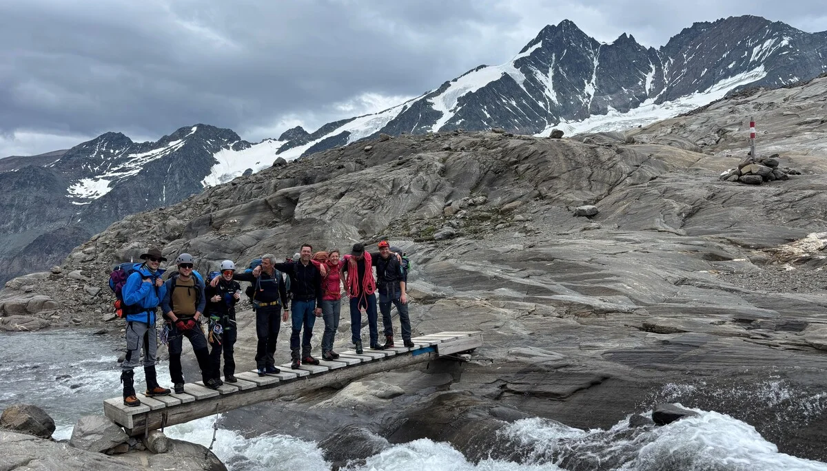 Sieben Bergsteiger mit Ausrüstung stehen auf einer schmalen Holzbrücke über einem Gebirgsbach, umgeben von Felsen und schneebedeckten Bergen. | © Steffen Wolf
