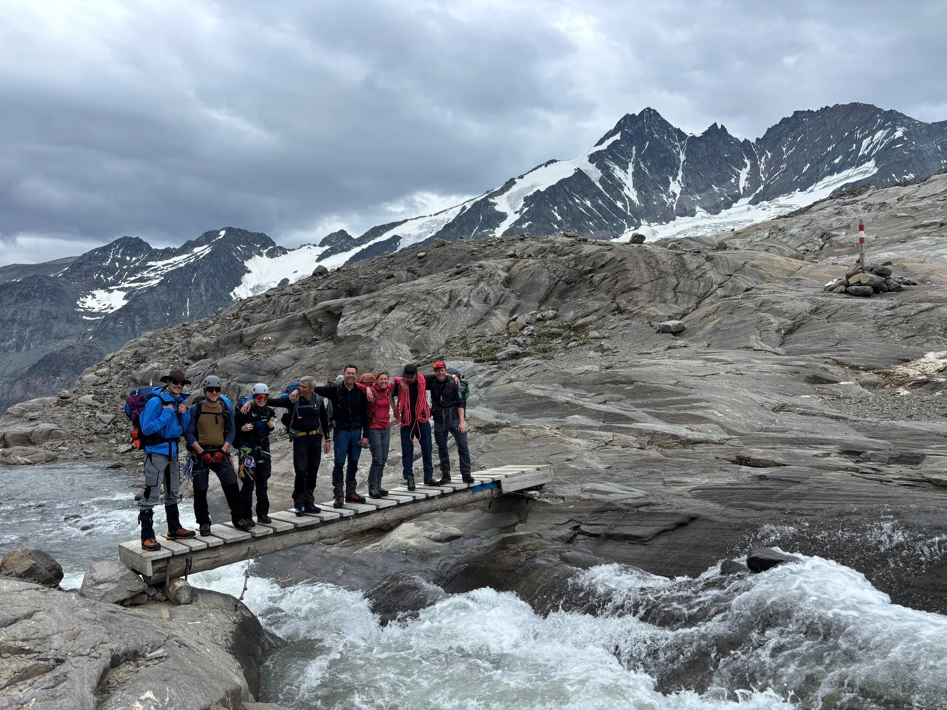 Sieben Bergsteiger mit Ausrüstung stehen auf einer schmalen Holzbrücke über einem Gebirgsbach, umgeben von Felsen und schneebedeckten Bergen. | © DAV Sektion Geltendorf