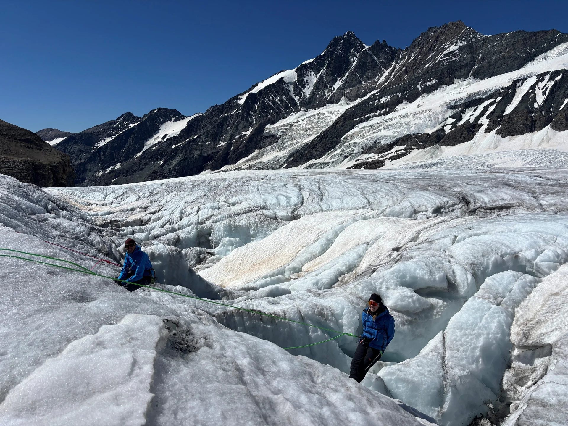 Zwei Personen in blauen Jacken auf einem Gletscher mit tiefen Spalten, umgeben von schneebedeckten Bergen | © Steffen Wolf