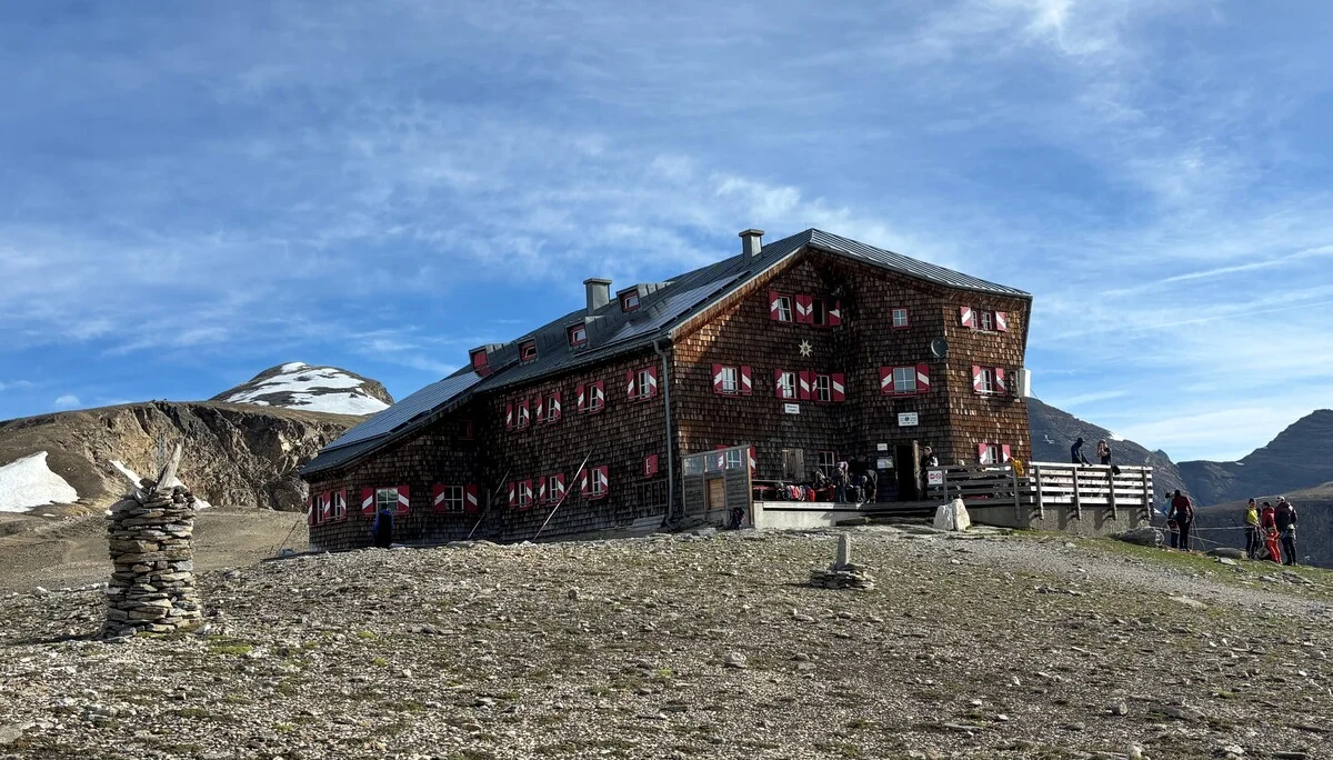 Berghütte mit rot-weiß gestrichenen Fensterläden auf felsigem Gelände unter blauem Himmel, im Hintergrund schneebedeckter Berg | © Steffen Wolf