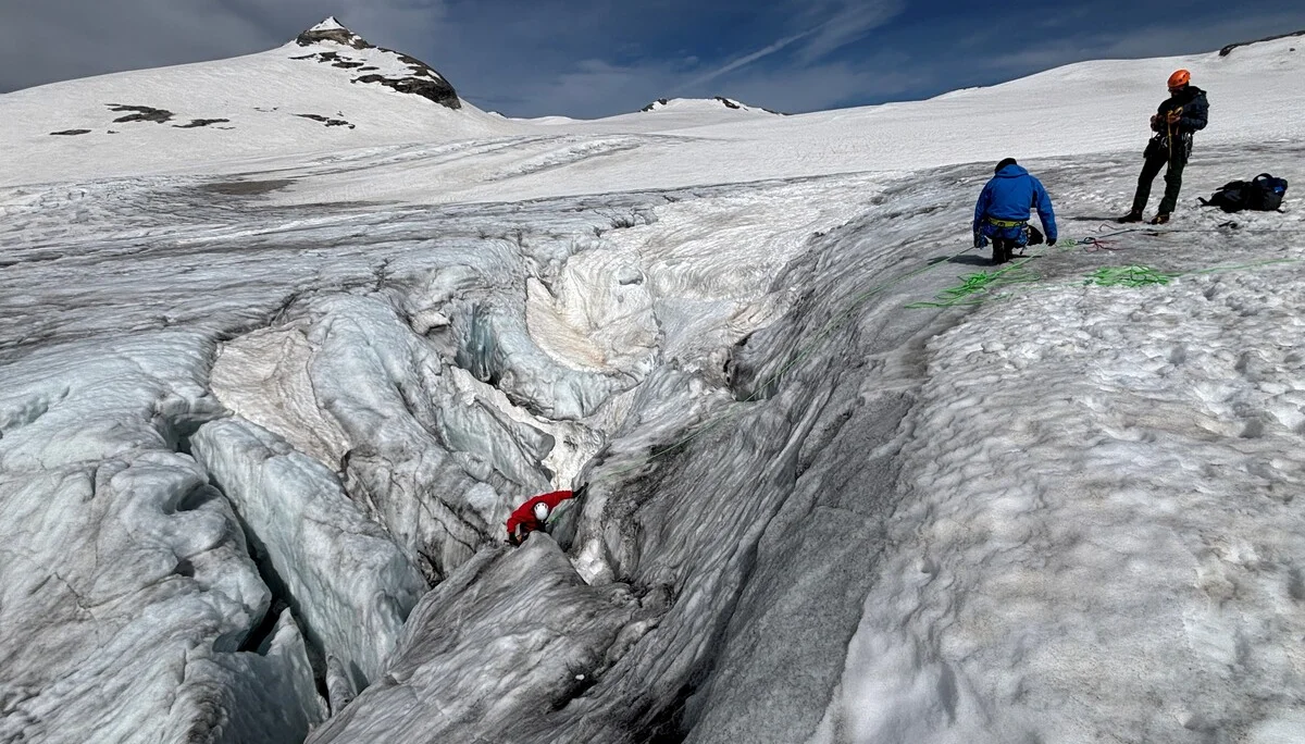 Zwei Personen in Bergsteigerausrüstung auf einem Gletscher neben einer tiefen Spalte, eine Person klettert hinein | © Steffen Wolf