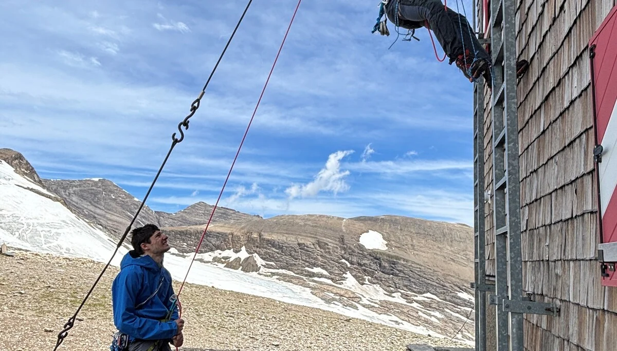 Eine Person klettert an einer Seilwand an einem Berghaus, eine andere sichert am Boden mit Kletterausrüstung, Berglandschaft im Hintergrund | © Steffen Wolf