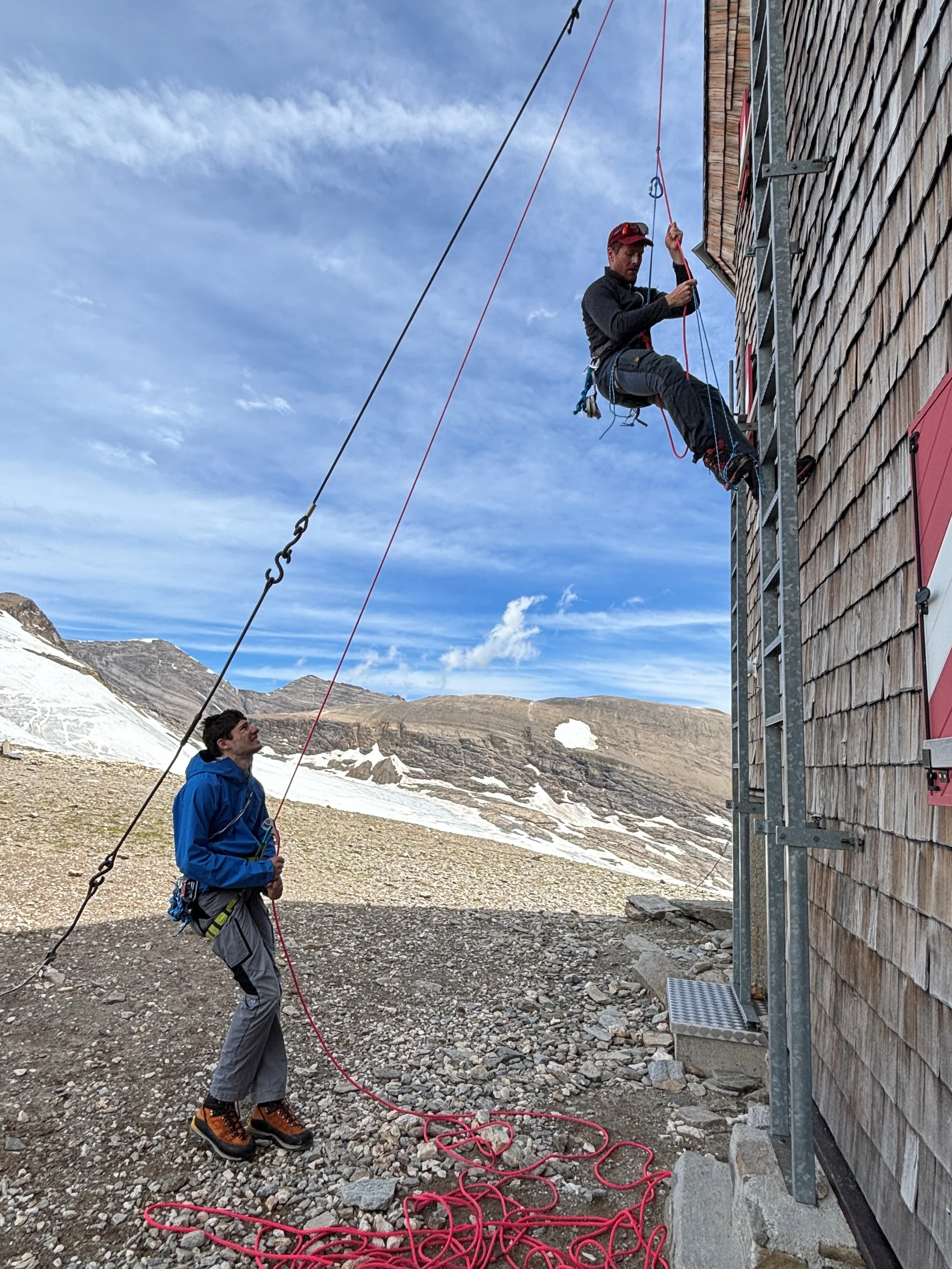 Eine Person klettert an einer Seilwand an einem Berghaus, eine andere sichert am Boden mit Kletterausrüstung, Berglandschaft im Hintergrund | © Steffen Wolf