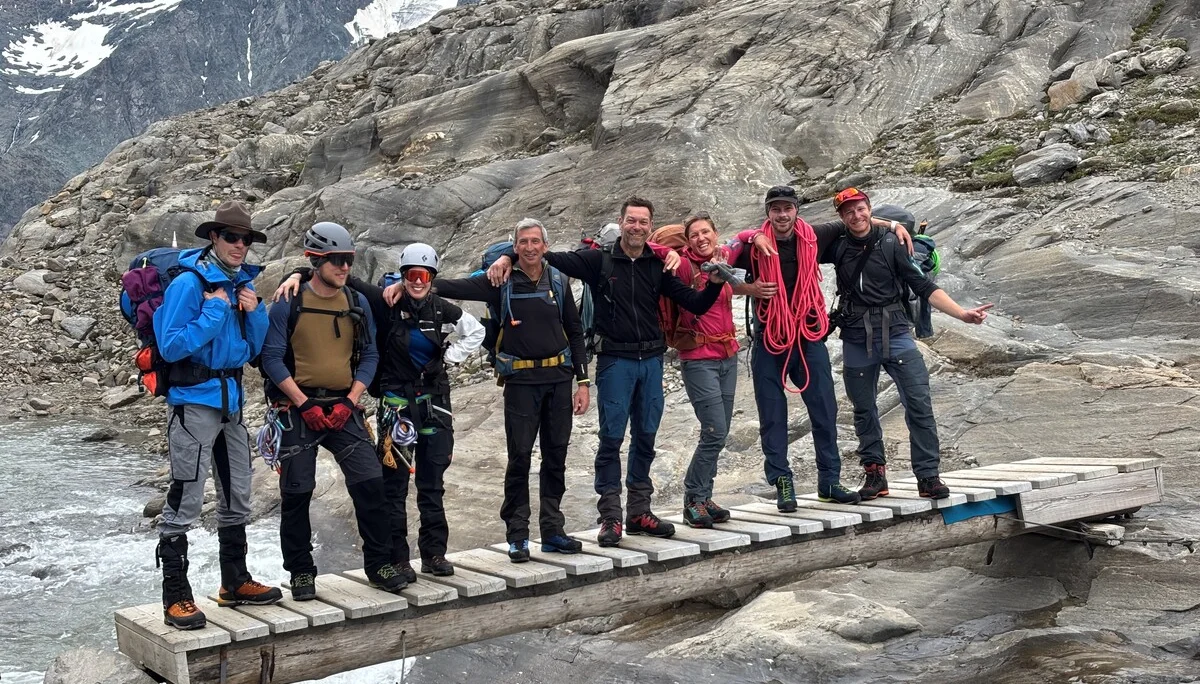 Sieben Bergsteiger mit Ausrüstung stehen auf einer schmalen Holzbrücke über einem Gebirgsbach, umgeben von Felsen und schneebedeckten Bergen. | © Steffen Wolf