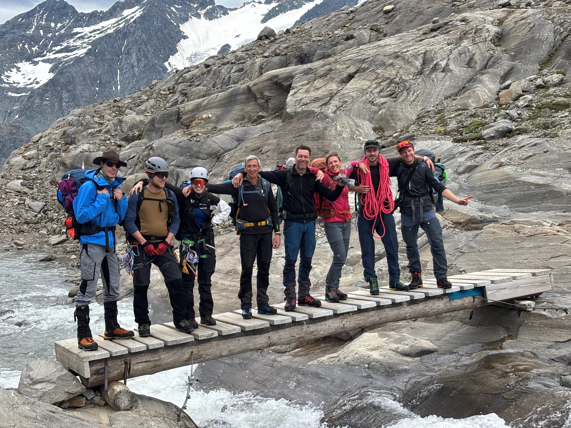 Sieben Bergsteiger mit Ausrüstung stehen auf einer schmalen Holzbrücke über einem Gebirgsbach, umgeben von Felsen und schneebedeckten Bergen. | © Steffen Wolf