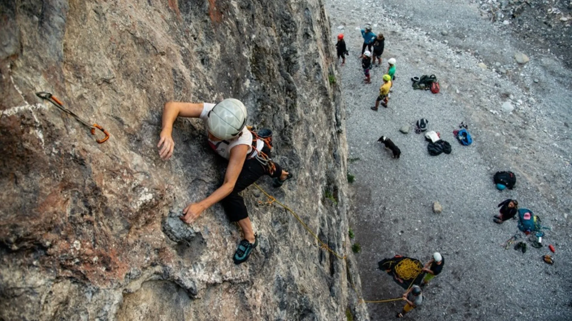 Kletterer mit Helm und Kletterausrüstung steigt an steiler Felswand, darunter Gruppe von Personen und Ausrüstung auf Geröllboden | © DAV Sektion Geltendorf