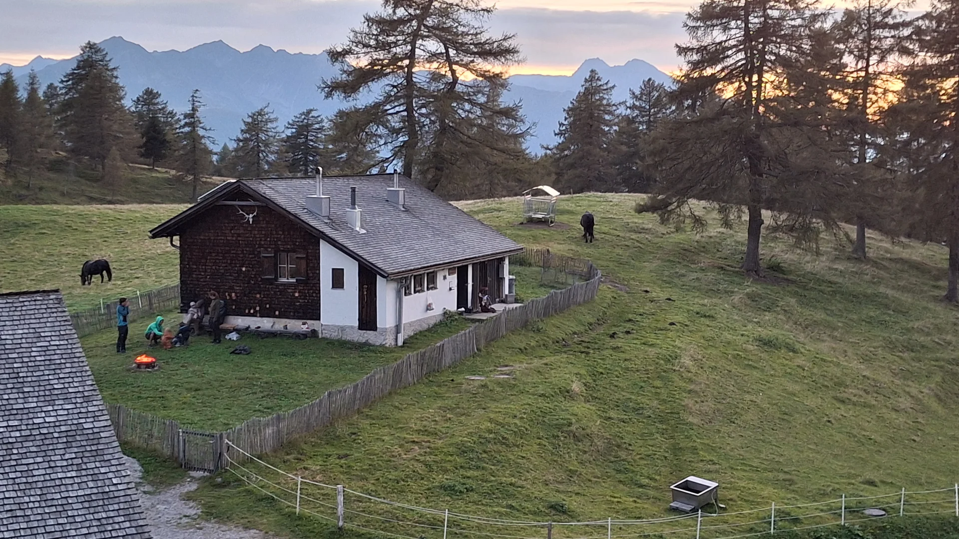 Jagdhaus auf einer grünen Wiese mit Zaun, Menschen um ein Lagerfeuer, zwei Pferde und Berge im Hintergrund bei Sonnenuntergang | © DAV Sektion Geltendorf