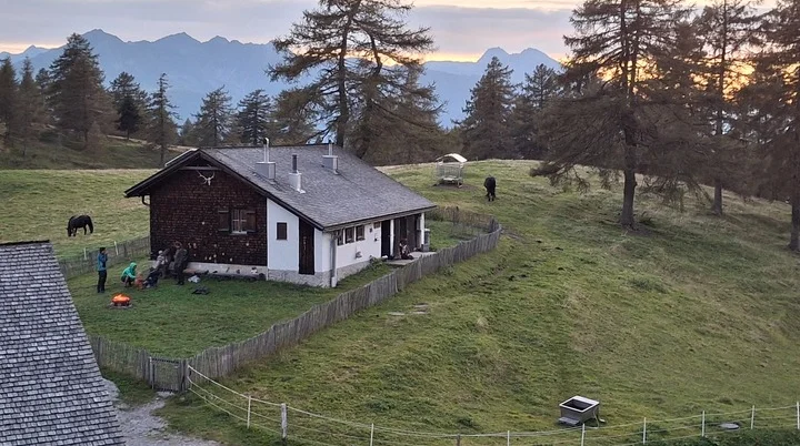 Jagdhaus auf einer grünen Wiese mit Zaun, Menschen um ein Lagerfeuer, zwei Pferde und Berge im Hintergrund bei Sonnenuntergang | © DAV Sektion Geltendorf