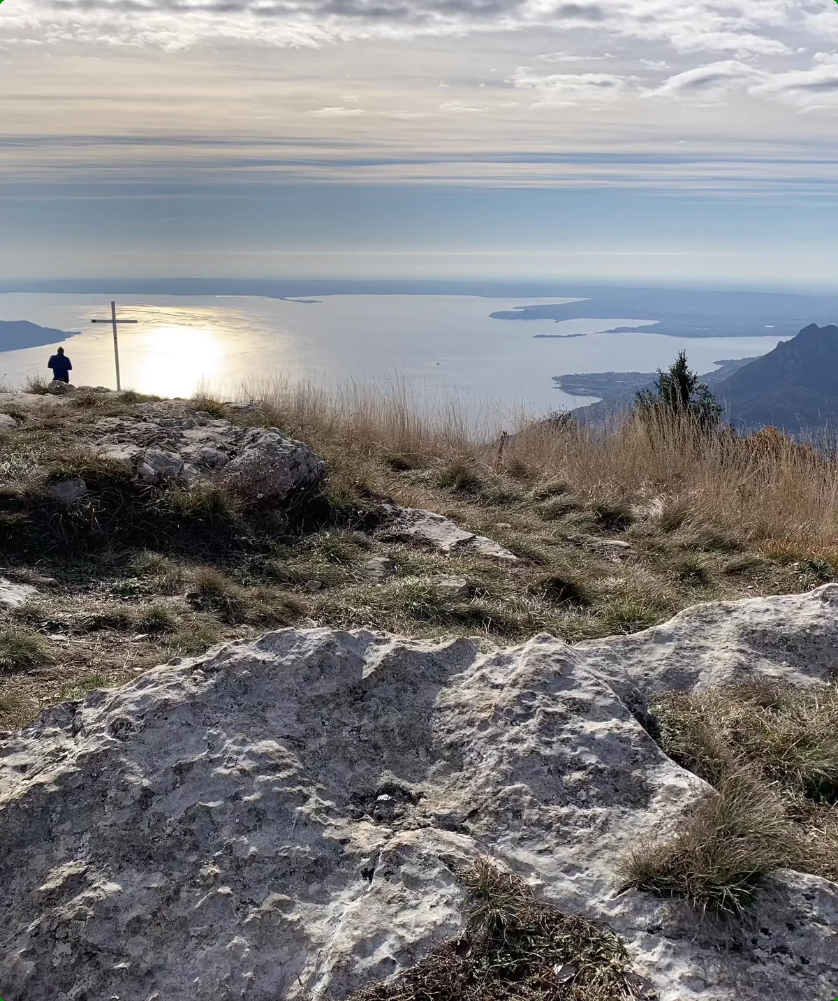 Berggipfel mit Felsen und Gras, Person sitzt neben einem Holzkreuz, Blick auf See und bewölkten Himmel | © DAV Sektion Geltendorf