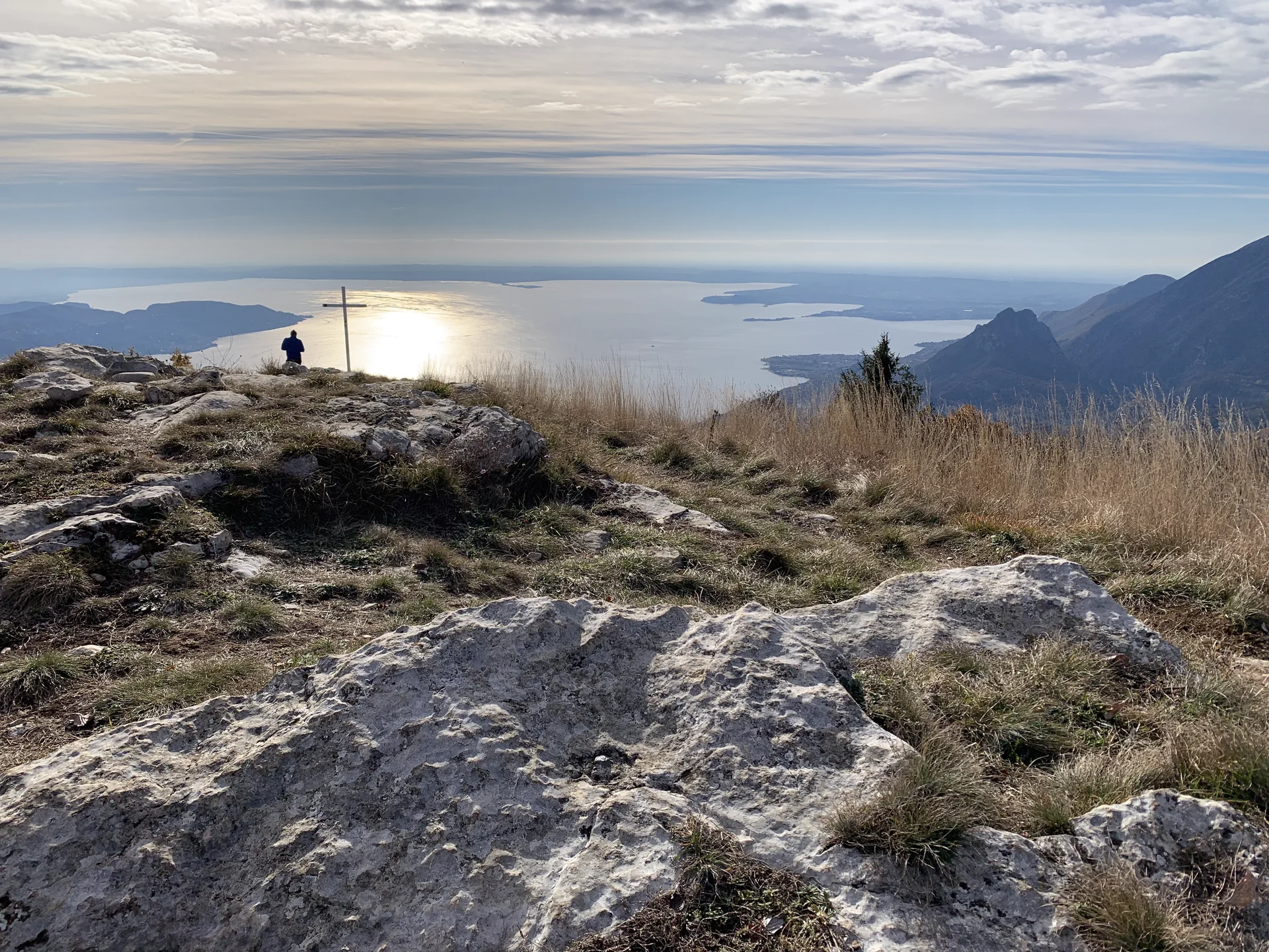 Berggipfel mit Felsen und Gras, Person sitzt neben einem Holzkreuz, Blick auf See und bewölkten Himmel | © DAV Sektion Geltendorf