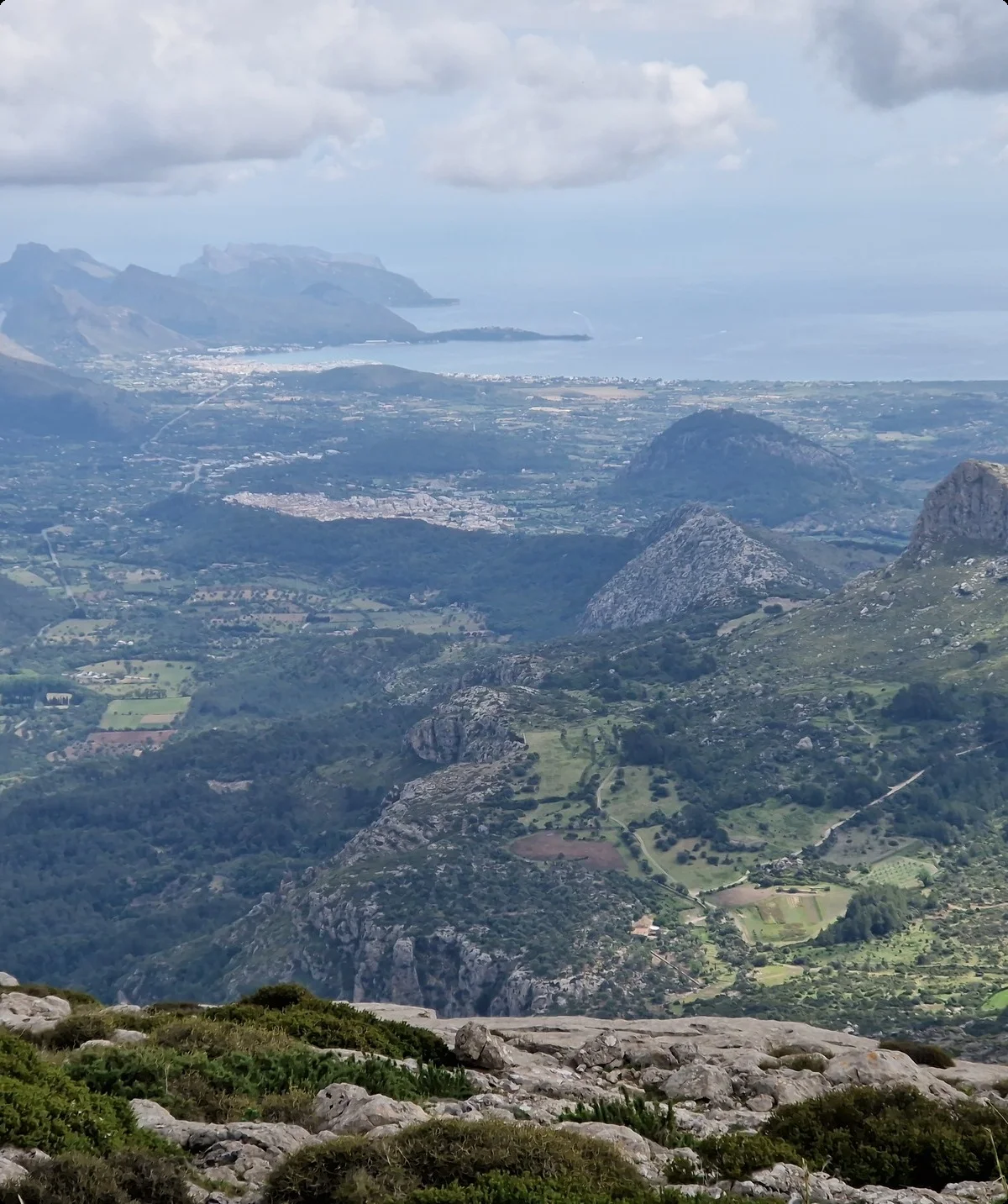 Weitläufige Aussicht von einem Berggipfel auf eine grüne, hügelige Landschaft mit Feldern, Wäldern und kleinen Siedlungen, im Hintergrund eine zerklüftete Küste und das Meer unter bewölktem Himmel. | © Nadine Daumann