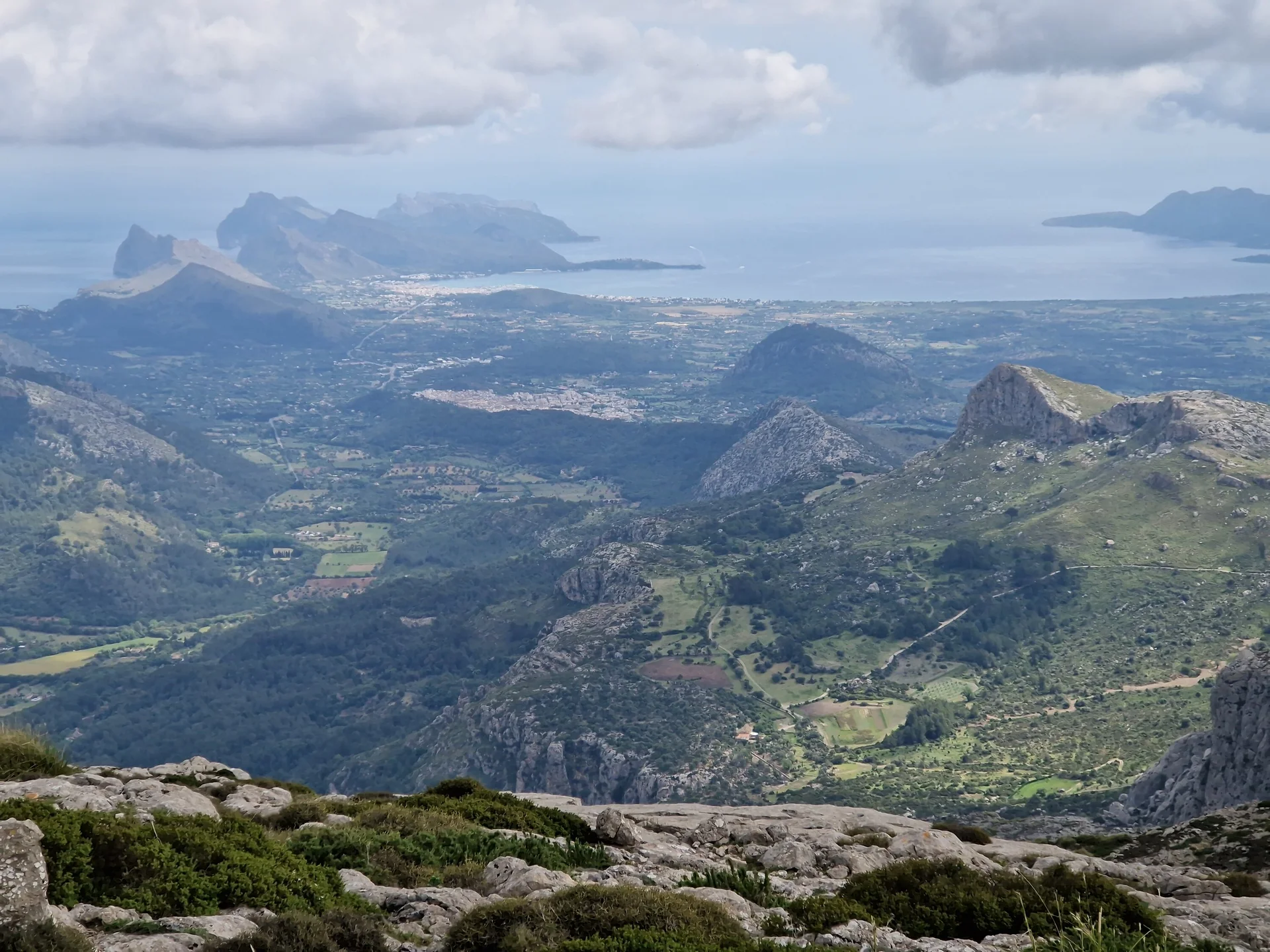 Weitläufige Aussicht von einem Berggipfel auf eine grüne, hügelige Landschaft mit Feldern, Wäldern und kleinen Siedlungen, im Hintergrund eine zerklüftete Küste und das Meer unter bewölktem Himmel. | © Nadine Daumann