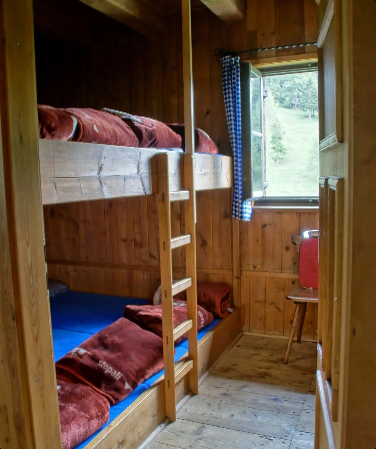 Schlafraum im Jägerhäusl mit zwei Etagenbetten aus Holz, roten Decken, offenem Fenster mit blau-weiß kariertem Vorhang und Blick auf eine grüne Wiese | © Sektion Geltendorf
