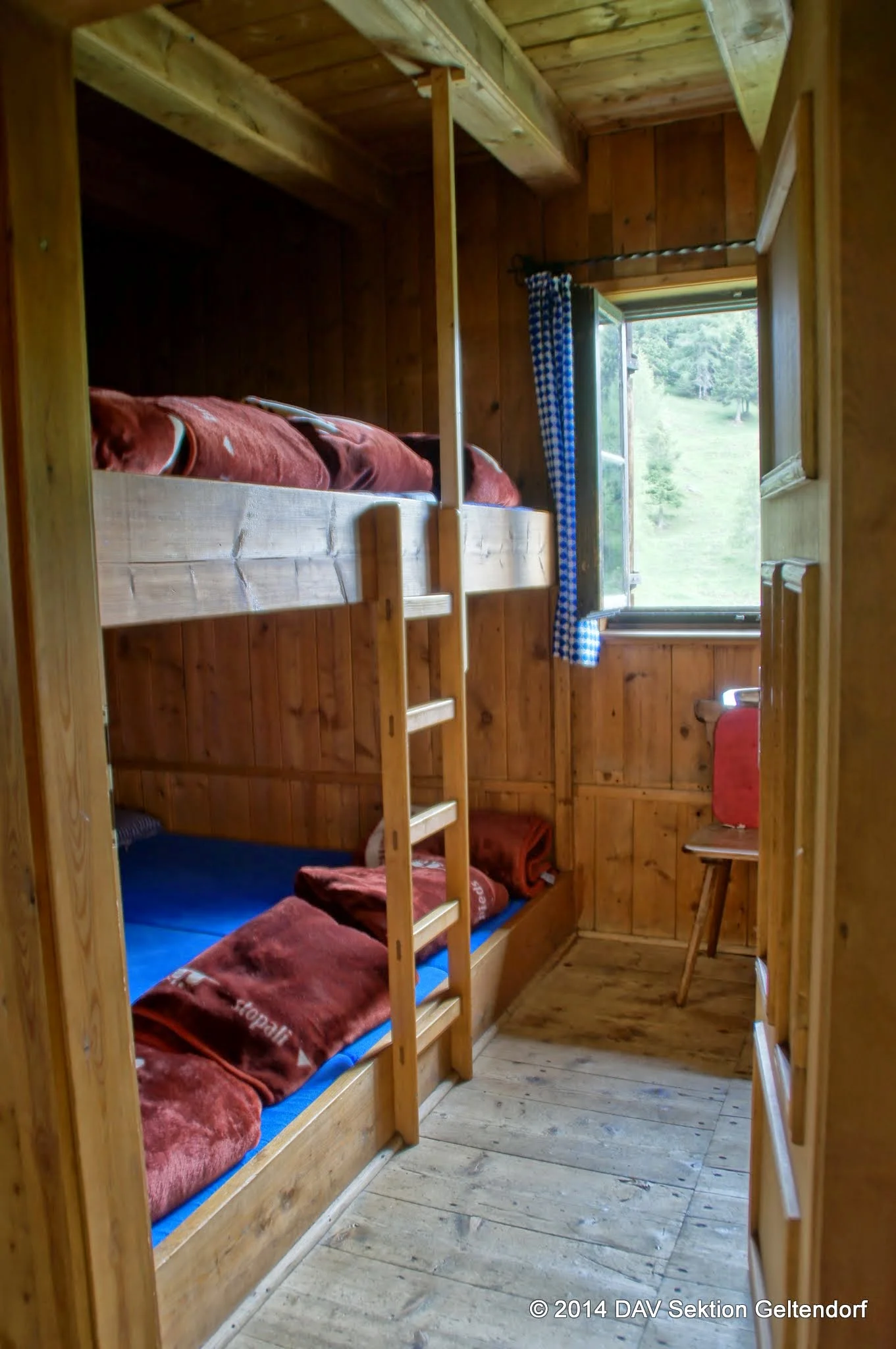 Schlafraum im Jägerhäusl mit zwei Etagenbetten aus Holz, roten Decken, offenem Fenster mit blau-weiß kariertem Vorhang und Blick auf eine grüne Wiese | © Sektion Geltendorf