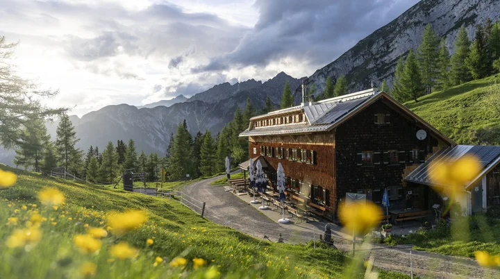 Neue Magdeburger Hütte, eine Berghütte in den Alpen, umgeben von Wiesen mit gelben Blumen und Bergen im Hintergrund unter bewölktem Himmel | © DAV/Wolfgang Ehn